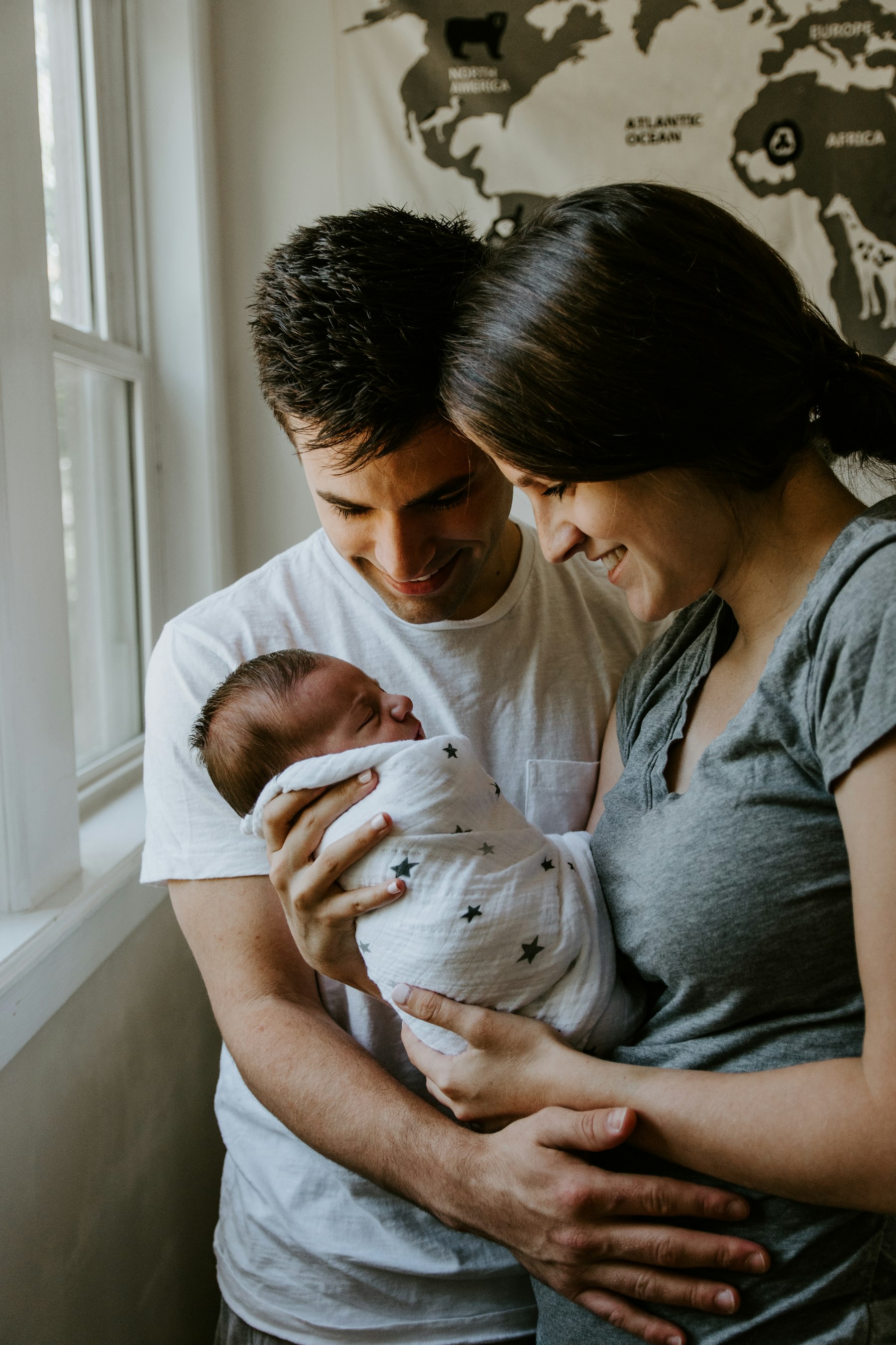 A couple at home by the window, smiling down at their newborn baby