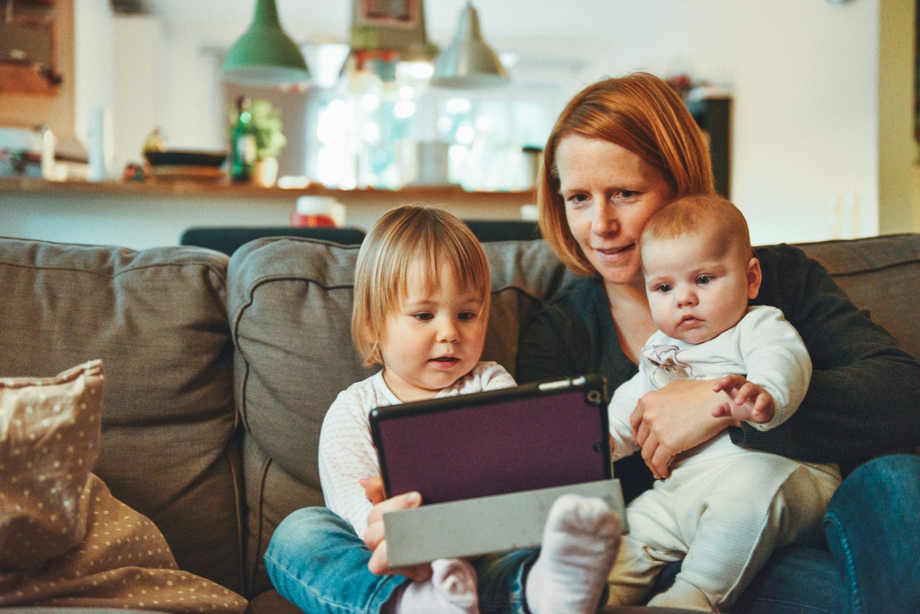 A mother at home on the sofa with her toddler and baby, looking at a tablet together