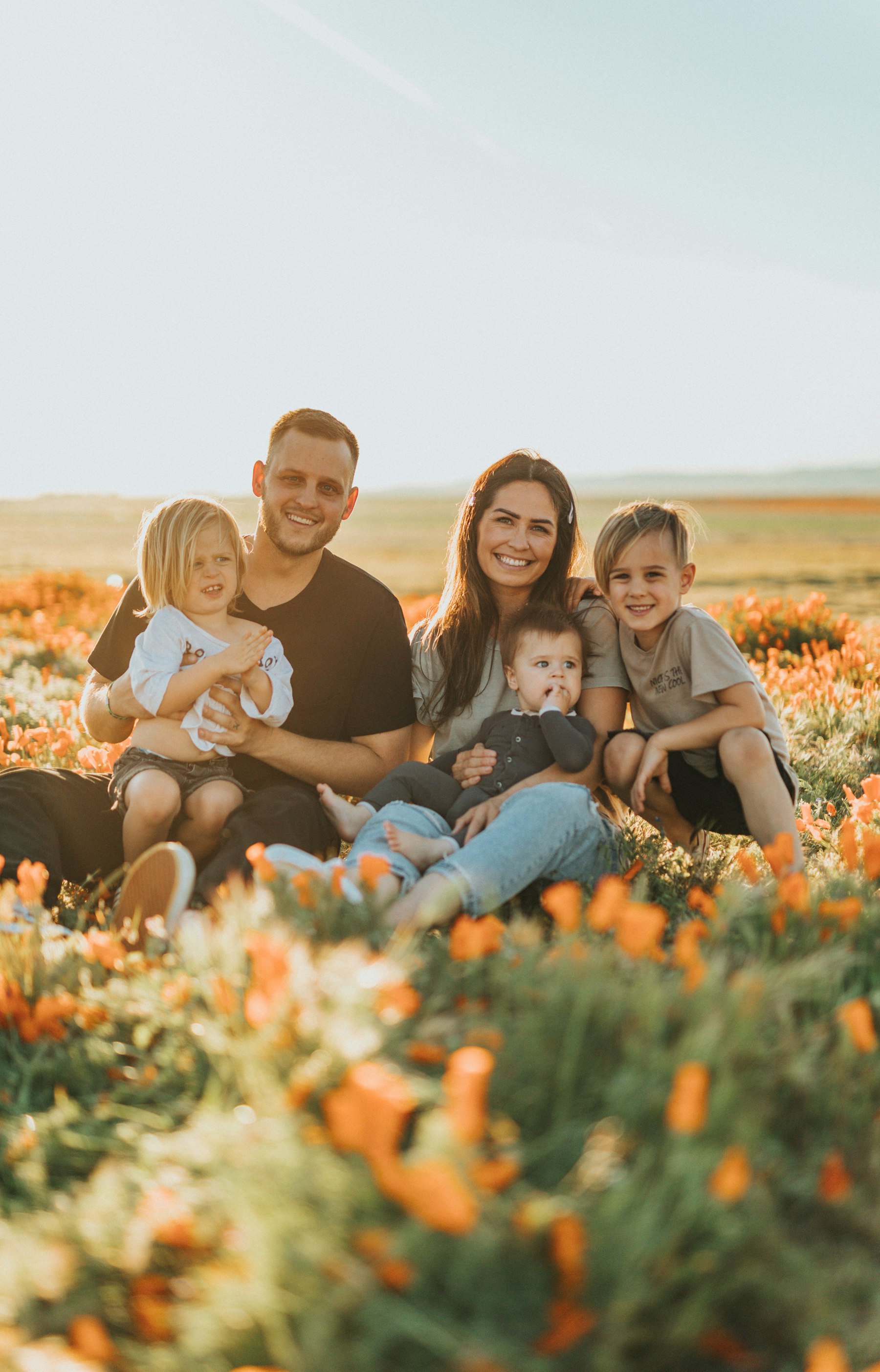 A smiling family of five sitting together in a sunlit meadow of wildflowers